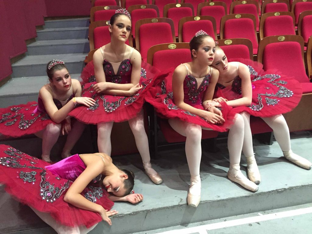 Image shows five young girls dressed in ballerina costumes at rest. Three girls have their eyes closed, one is lying down. Two of the girls are resting against each other. Photo by Tania Savona.