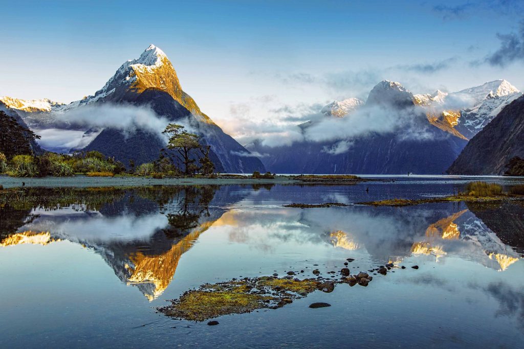 Image shows the water of Milford Sound in the foreground, with snow-capped mountains in the background. The mountains are reflected in the smooth surface of the water. Photo Kerry Boytell